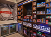 Snack shop interior with wooden shelves displaying chips, cookies, and assorted snacks. Refrigerated section below stocks drinks. A sign labeled "TREATS" is on the wall.