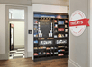 Hotel snack area with shelving displaying various packaged snacks and drinks, a small chalkboard with a welcome message, and a door leading to another room. A round sign reads "TREATS.
