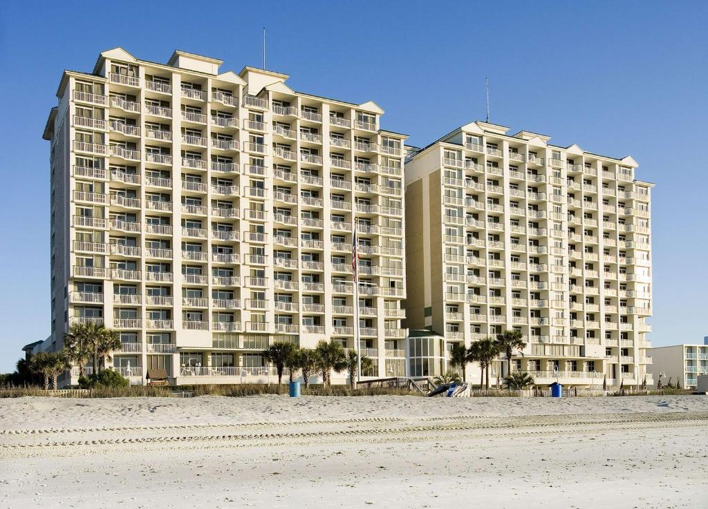 Two tall, beige beachfront condominium buildings with balconies, seen from the sandy beach under a clear blue sky. Small palm trees and beach amenities are visible in the foreground.