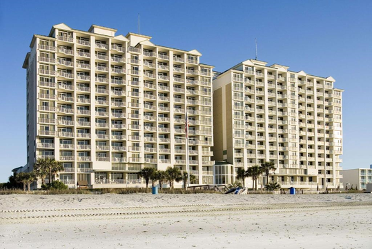 Two tall, beige beachfront condominium buildings with balconies, seen from the sandy beach under a clear blue sky. Small palm trees and beach amenities are visible in the foreground.