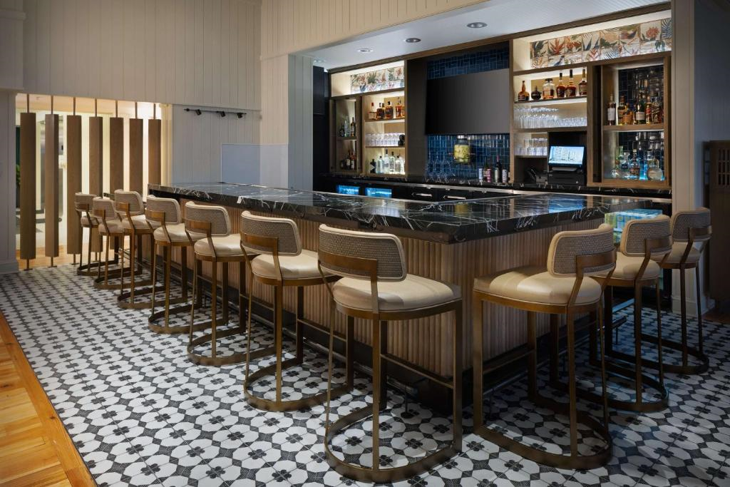 A modern bar with a marble countertop, cushioned barstools, patterned tile floor, shelves of liquor bottles, and a blank screen behind the counter.