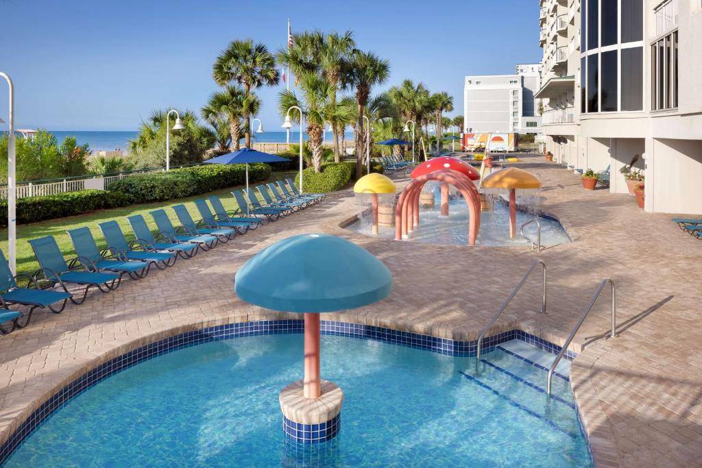 Outdoor pool area with mushroom-shaped water features, lounge chairs, palm trees, and a view of the ocean in the background.