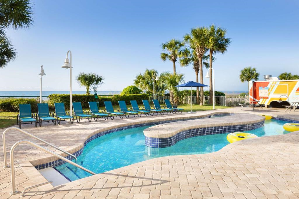 Outdoor swimming pool with lounge chairs, palm trees, and a view of the ocean in the background under a clear blue sky.