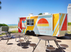 A colorful food truck labeled "Golden Hour Drinking Deck" is parked by outdoor tables on a sunny day near the beach.