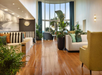 Modern hotel lobby with polished wooden floor, striped and neutral sofas, green plants, large windows, and natural light streaming in.