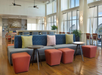 Modern lounge area with a blue and gray sofa, colorful cushions, orange ottomans, small round tables, wooden floor, and large windows letting in natural light.