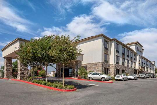 A three-story hotel building with a stone facade and covered entrance, surrounded by parked cars and landscaped trees under a partly cloudy sky.
