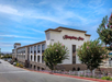 Exterior view of a Hampton Inn hotel with a beige facade, red signage, multiple windows, and trees in front.
