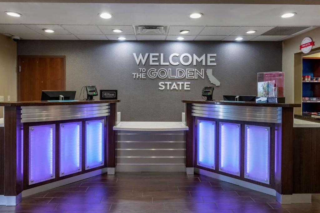 Hotel front desk area with a modern design featuring illuminated panels. A sign reads "Welcome to the Golden State." Two computer terminals are on the counter.