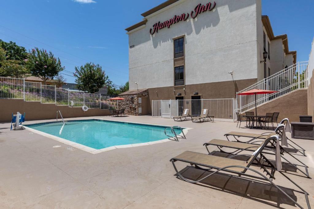 Outdoor hotel pool area with lounge chairs and umbrellas beside a fenced pool. A multi-story building is in the background.