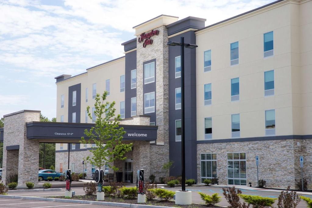 Four-story hotel building with stone and stucco exterior, signage for Hampton Inn. Foreground includes a parking lot with EV charging stations and landscaped areas.