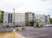 Four-story hotel with a modern facade, flanked by parked cars, with flags at the entrance and a spacious parking lot in front.