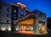 Night view of a Hampton Inn entrance with illuminated signage. The building features stone facades and a covered drop-off area, surrounded by landscaping and lit by exterior lights.
