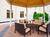 Outdoor patio area with a wooden gazebo, brick flooring, and a set of wicker furniture with beige cushions. The background shows a light-colored building and green shrubs.