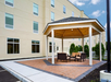 Outdoor seating area with a wooden gazebo, chairs, and tables on a brick patio, adjacent to a beige multi-story building.