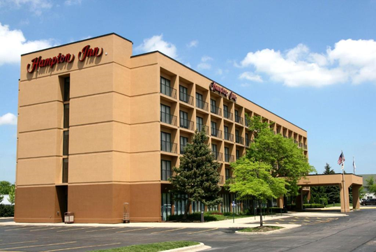 Four-story Hampton Inn hotel with beige exterior, red signage, and surrounding trees, under a blue sky with clouds. Blacktop parking lot in front.