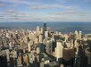 Aerial view of a city skyline with numerous skyscrapers under a partly cloudy sky, adjacent to a large body of water.