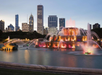 Colorful, illuminated fountain in a park with a city skyline in the background at dusk.
