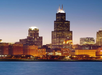 Night view of the Chicago skyline, featuring illuminated skyscrapers, including the Willis Tower, with a calm body of water in the foreground.