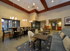 Hotel lounge area with high tables, cushioned chairs, a large sofa, and pendant lighting. The room features a modern design with neutral tones and a patterned rug.