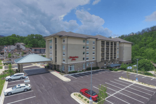 Aerial view of a Hampton Inn hotel with a large parking lot and surrounding greenery under a cloudy sky.
