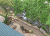 Aerial view of a landscaped backyard featuring a patio with chairs, a fire pit, large rocks, and greenery alongside a flowing creek.