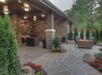 Covered patio with stone flooring, wicker furniture, and a fire pit. Tall shrubs and plants are arranged around the space.