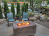 A patio with a rectangular fire pit, two chairs, and surrounding greenery, near a stream with rocks.