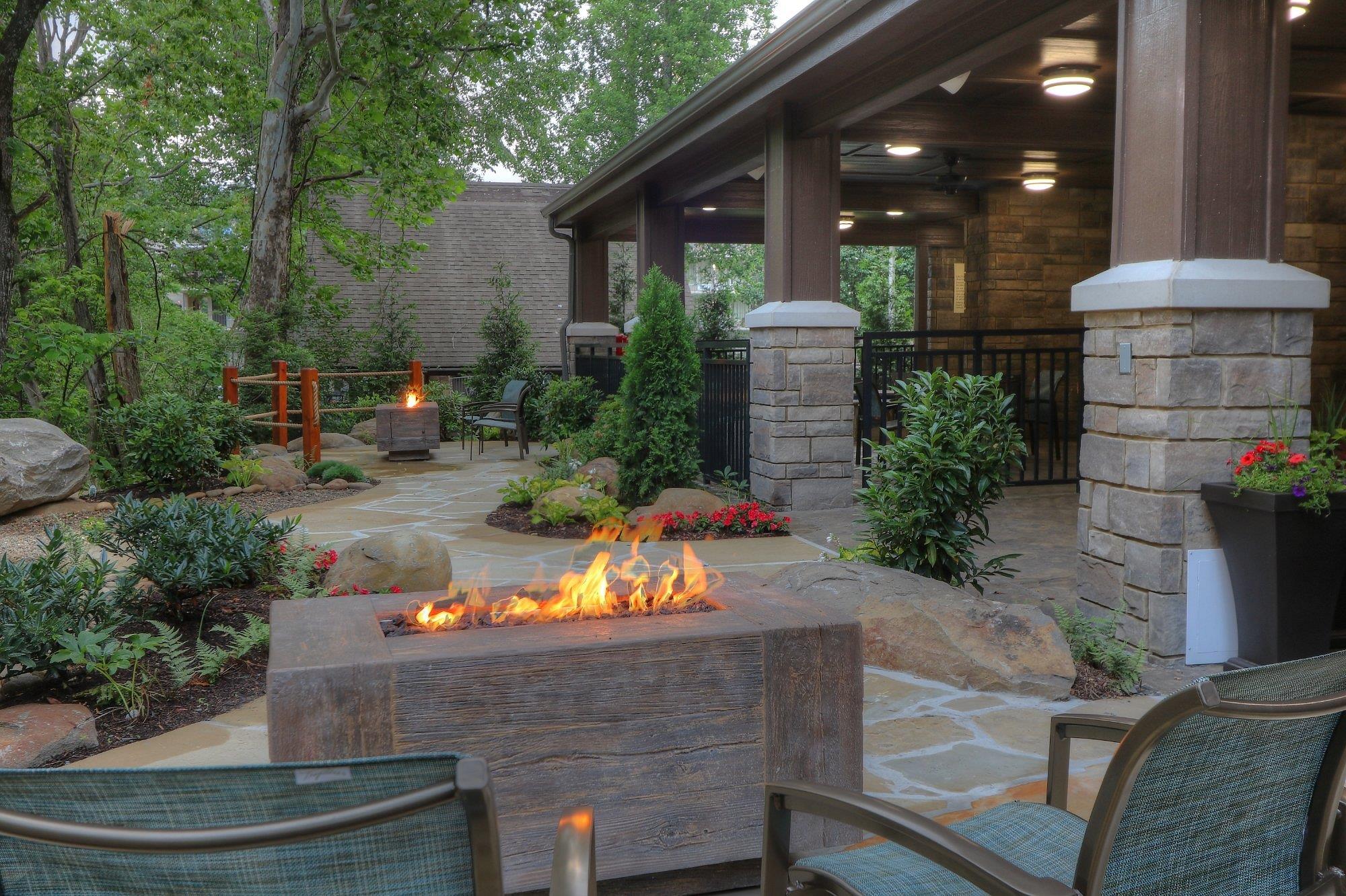 A modern patio with chairs surrounds a rectangular gas fire pit, with landscaped plants and a covered seating area in the background.