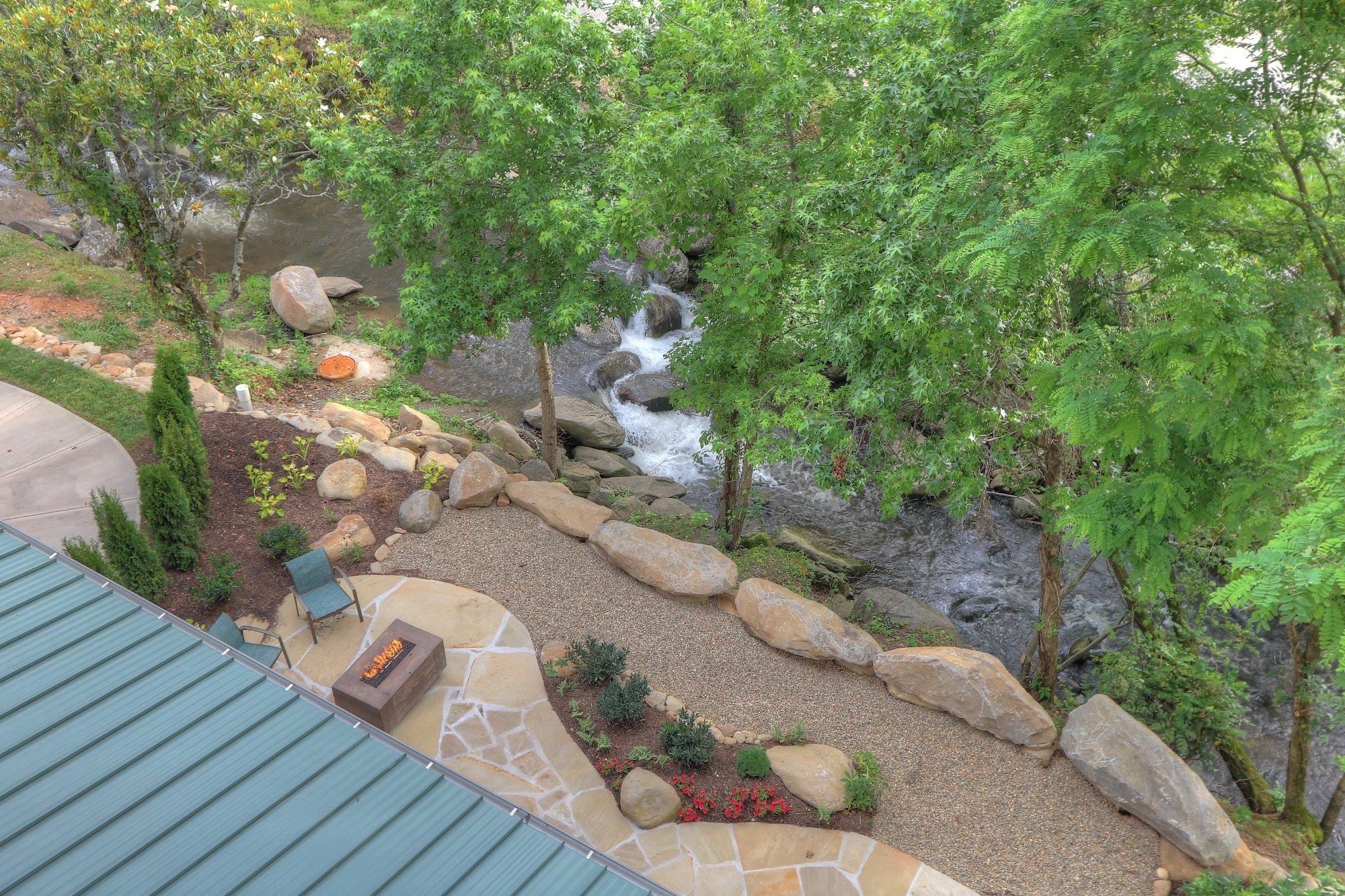 Aerial view of a landscaped patio with chairs and a firepit beside a creek, surrounded by trees, rocks, and a metal-roofed building.