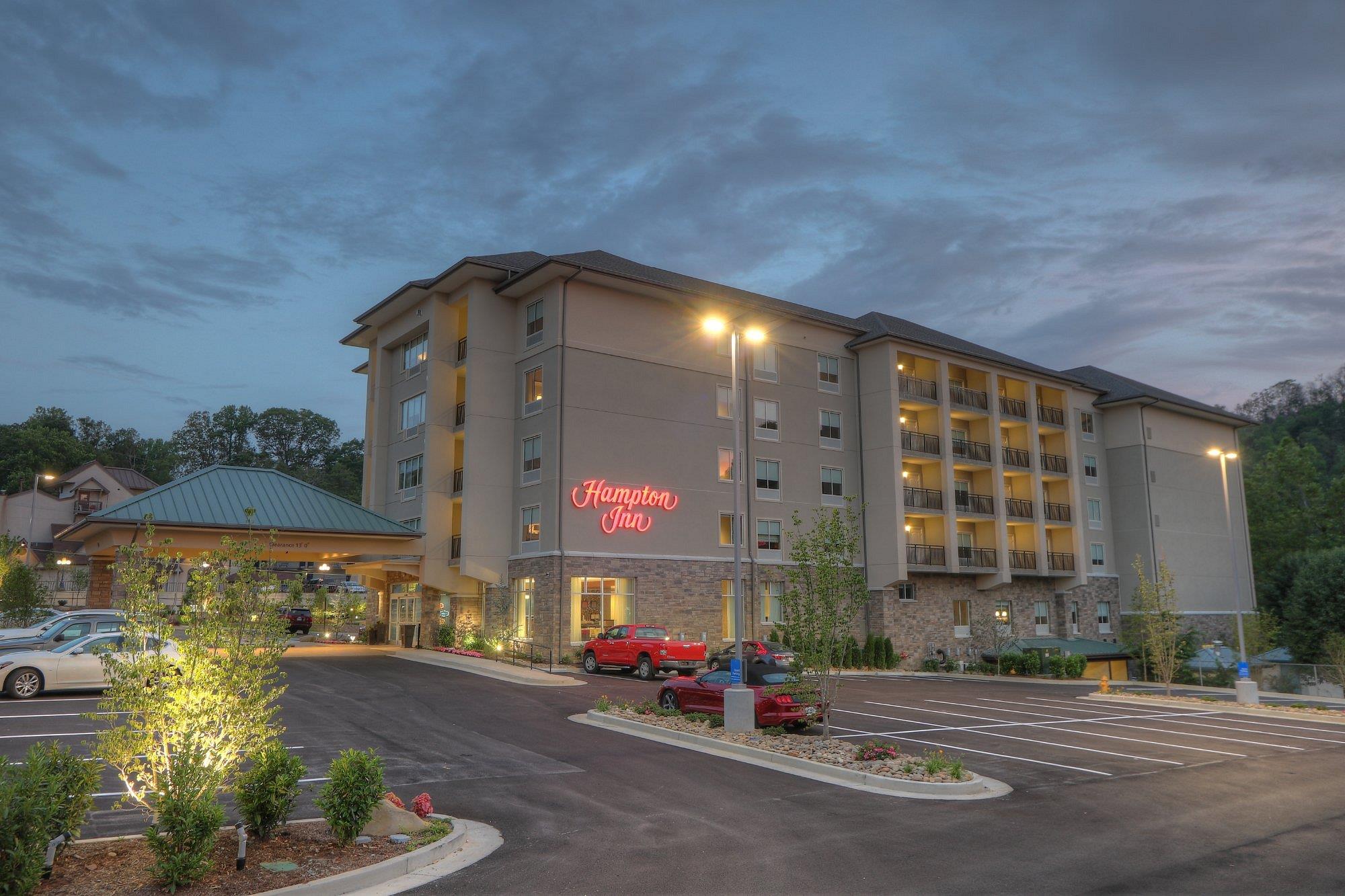 Exterior view of a Hampton Inn hotel at dusk, with cars parked in the lot, illuminated signs, and landscaped surroundings.