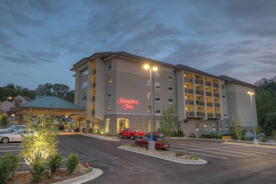 Exterior view of a Hampton Inn hotel at dusk, with cars parked in the lot, illuminated signs, and landscaped surroundings.