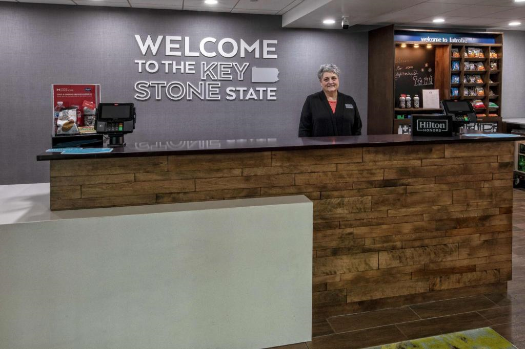 Hotel front desk with a staff member standing behind a wood-accented counter. A sign reads "Welcome to the Keystone State." Snack area visible in the background.