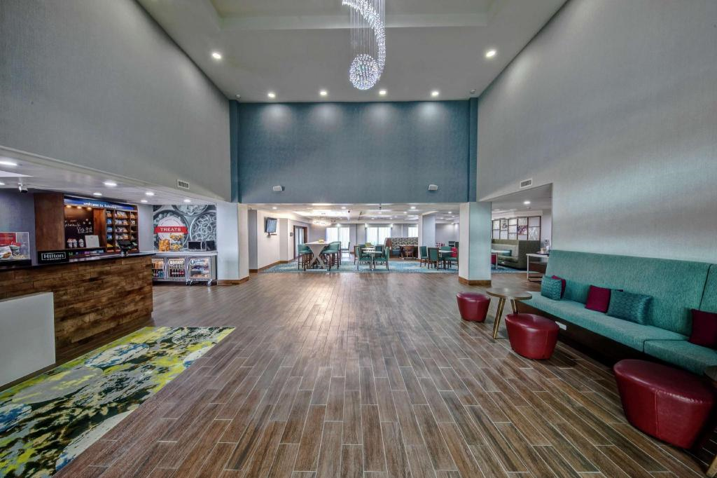 Spacious hotel lobby with wood flooring, a seating area, colorful rugs, and a modern chandelier. A reception desk and entrance to a dining area are visible.