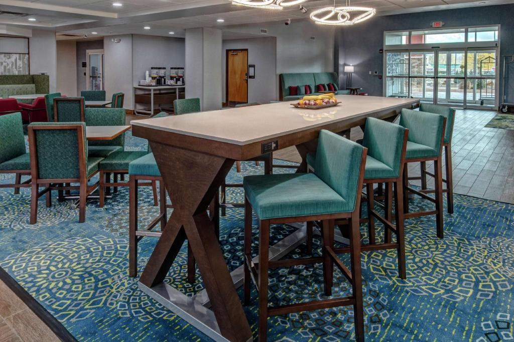 Hotel lobby featuring a tall table with green upholstered chairs, a patterned blue carpet, and modern lighting. Entrance doors are in the background.