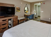 Hotel room interior featuring a bed, flat-screen TV on a wooden stand, desk with chair, wall art, and a doorway leading to another room.