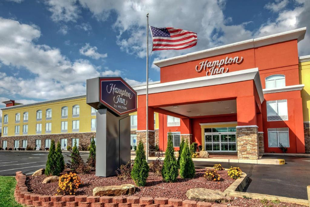 Hampton Inn hotel exterior with a prominent sign and American flag in front, surrounded by a landscaped area with small shrubs and plants, and a clear blue sky in the background.