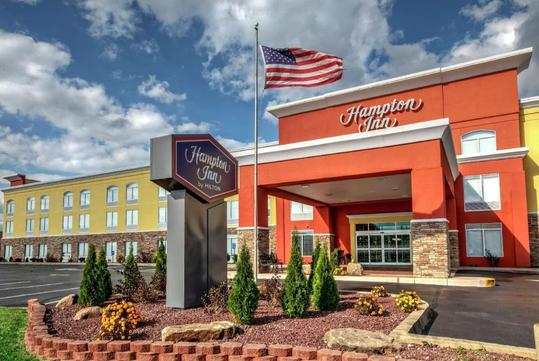 Hampton Inn hotel exterior with a prominent sign and American flag in front, surrounded by a landscaped area with small shrubs and plants, and a clear blue sky in the background.
