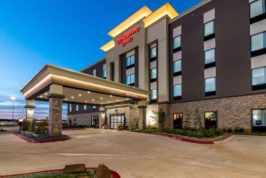 Exterior view of a Hampton Inn hotel at dusk, featuring stone accents, lit entrance, and multiple windows.