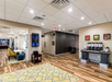 Hotel lobby with wood flooring, a seating area with tables and chairs, a coffee station, and framed art on cream-colored walls. Ceiling lights illuminate the space.