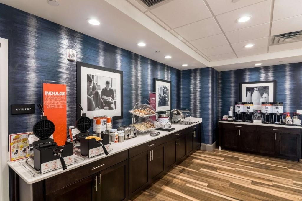 Breakfast buffet area with waffle makers, cereal dispensers, pastries, and coffee machines on a counter, against a blue textured wall with framed black and white photos.
