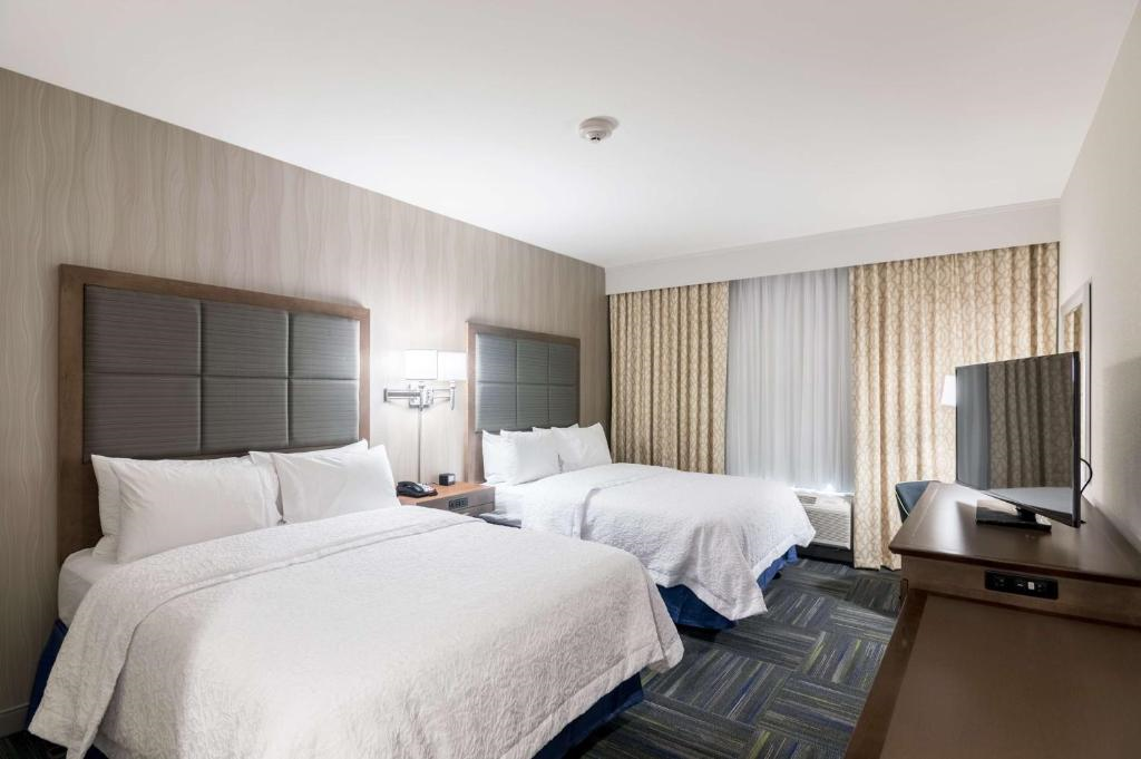 Hotel room with two double beds, wall-mounted lamps, a flat-screen TV on a dresser, and a window with patterned curtains. Walls are light-colored, and the carpet is patterned in shades of blue and green.