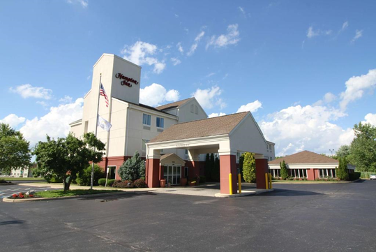 Exterior view of a Hampton Inn hotel with a clear blue sky, an American flag, and a spacious parking area in the foreground.
