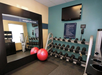 Hotel fitness room with a mirror, two exercise balls, a rack of dumbbells, a wall-mounted TV, and towels.