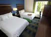 Hotel room with two double beds, white bedding, and a green and blue carpet. A window with green curtains is on the right, letting in natural light. A desk and chair are next to the window.