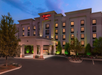 Exterior view of a Hampton Inn hotel at dusk, with illuminated windows and a red neon sign, surrounded by trees and a well-lit driveway.