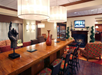 Hotel lobby with a long wooden table, modern decor, a sculpture of a hand, and a fireplace. A TV is mounted above the fireplace, and there are upholstered chairs around the table.