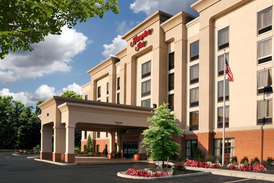 A four-story hotel building with a covered entrance, surrounded by landscaping. An American flag is on a pole to the right.