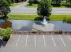 Aerial view of an empty parking lot beside a pond with a fountain, bordered by a grassy area and trees.