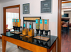 Continental breakfast station with cereal and milk dispensers on a countertop. Framed signs on the wall read: "Wake Up & Shine Happy" and "Bake the World a Better Place.